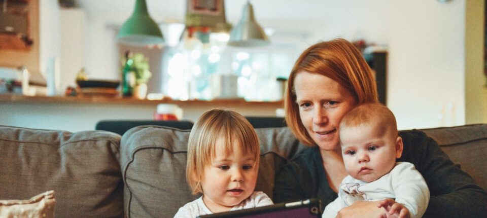 two babies and woman sitting on sofa while holding baby and watching on tablet